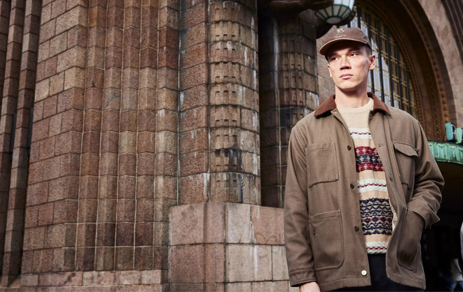 Man wearing a brown jacket and patterned sweater standing in front of the central railway station in Helsinki